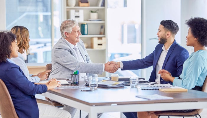 Two credit unions are merging and two executives in suits are shaking hands discussing the merger communication strategy.