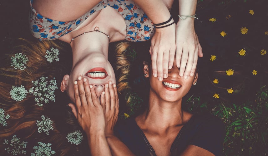 Two millennial girls covering each others eyes laughing.